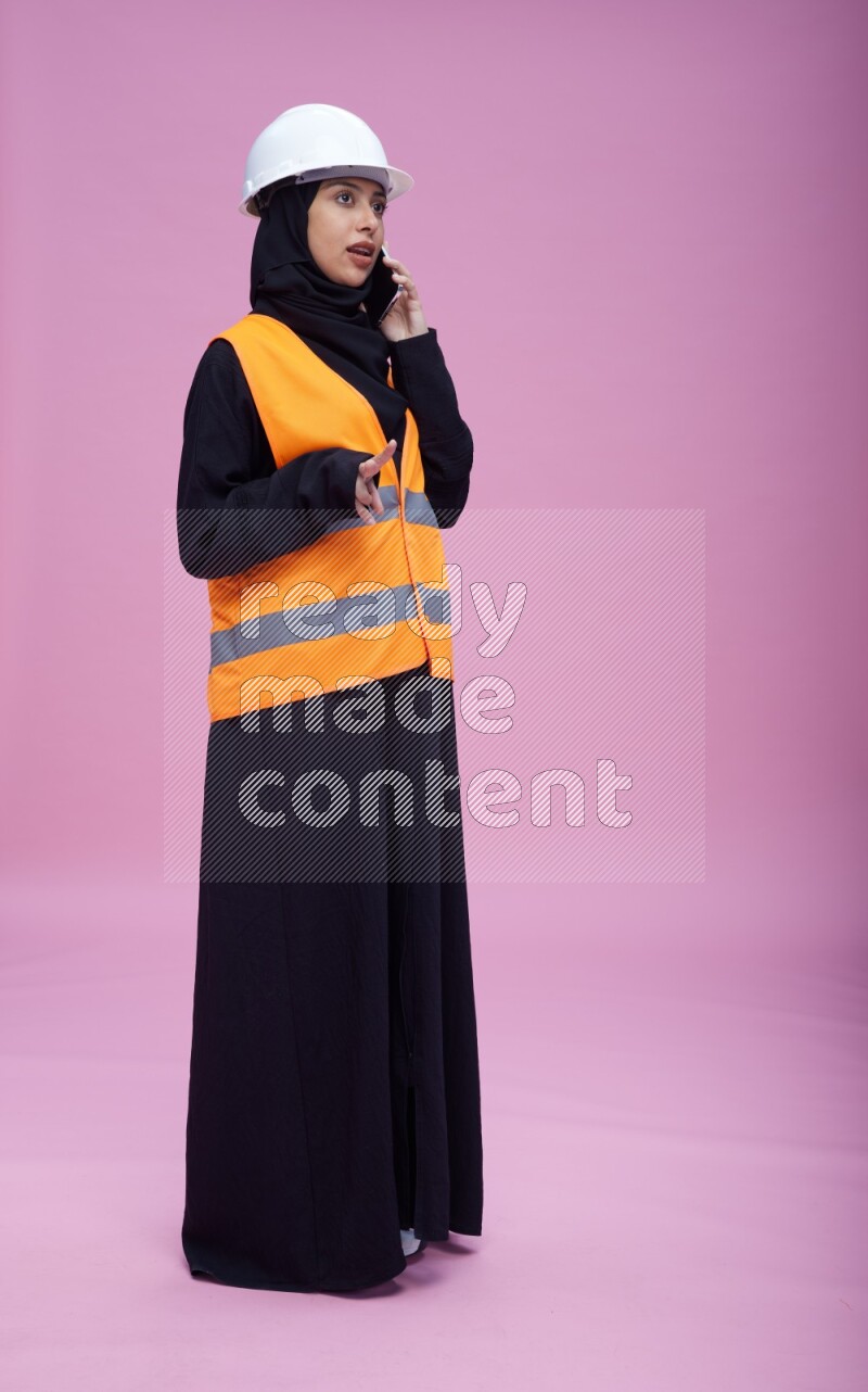 Saudi woman wearing Abaya with engineer vest and helmet standing talking on phone on pink background