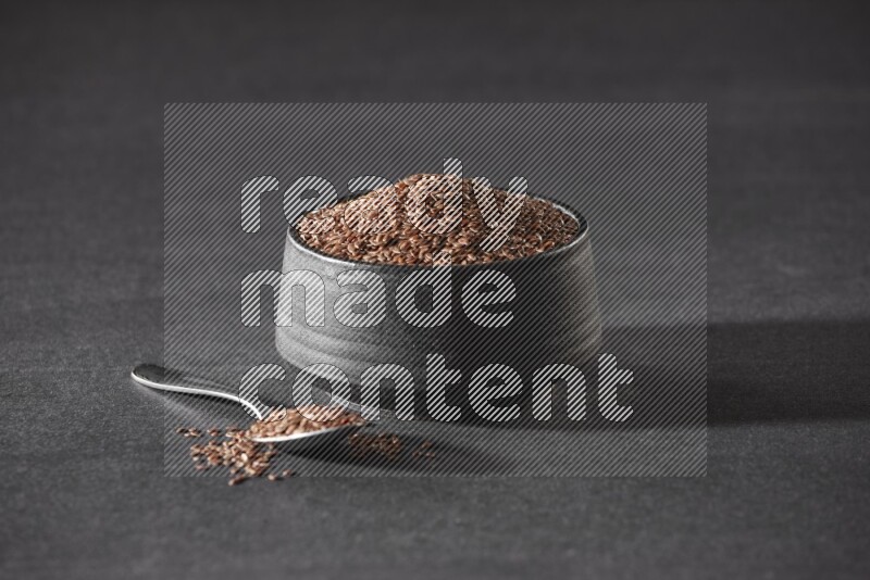 A black pottery bowl full of flaxseeds and a metal spoon full of the seeds on a black flooring