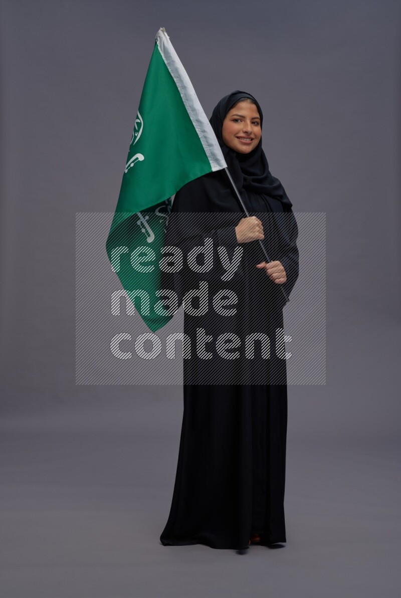 Saudi woman wearing Abaya standing holding Saudi flag on gray background