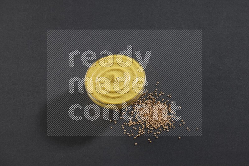 A glass bowl full of mustard paste with mustard seeds underneath on black flooring