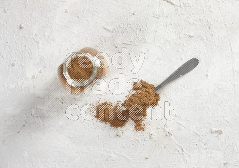 Herbs glass jar full of cinnamon powder with a metal spoon full of powder on a textured white background