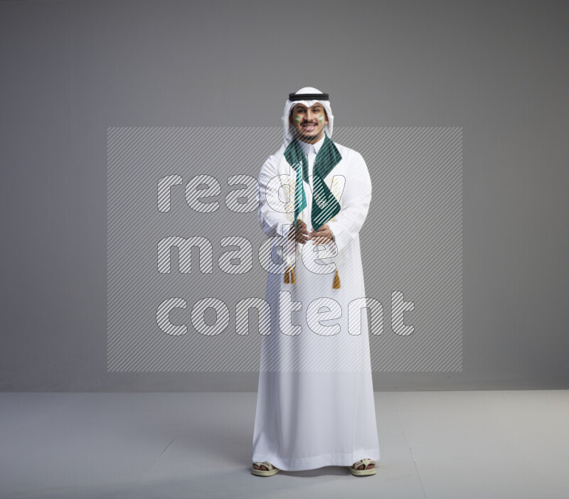 A saudi man standing wearing thob and white shomag with face painting and saudi flag scarf and holding small saudi flag on gray background