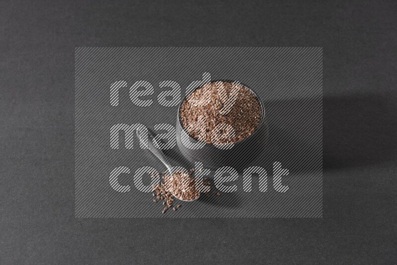A black pottery bowl full of flaxseeds and a metal spoon full of the seeds on a black flooring