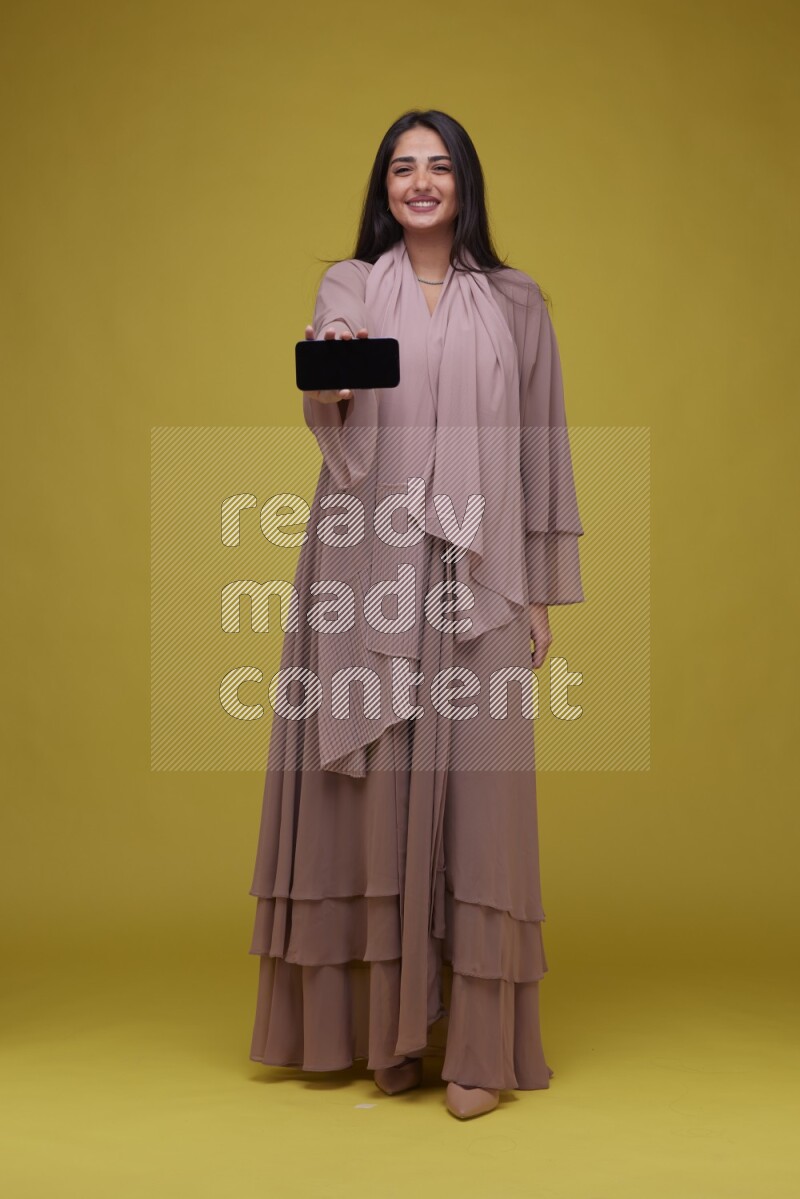 A woman Showing SmartPhone Screen on a Yellow Background wearing Brown Abaya