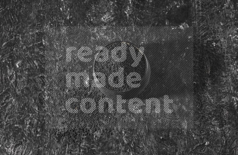 A black pottery bowl full of black seeds on a textured black flooring