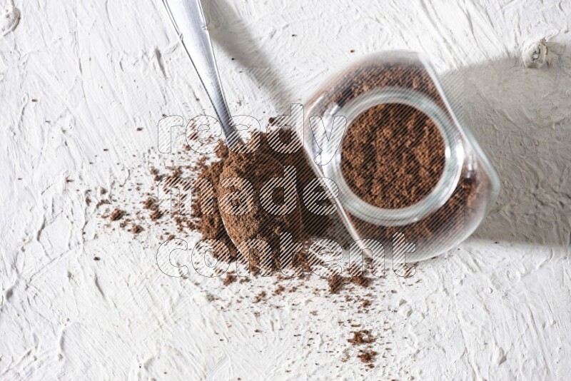 A glass spice jar and a metal spoon full of cloves powder on textured white flooring