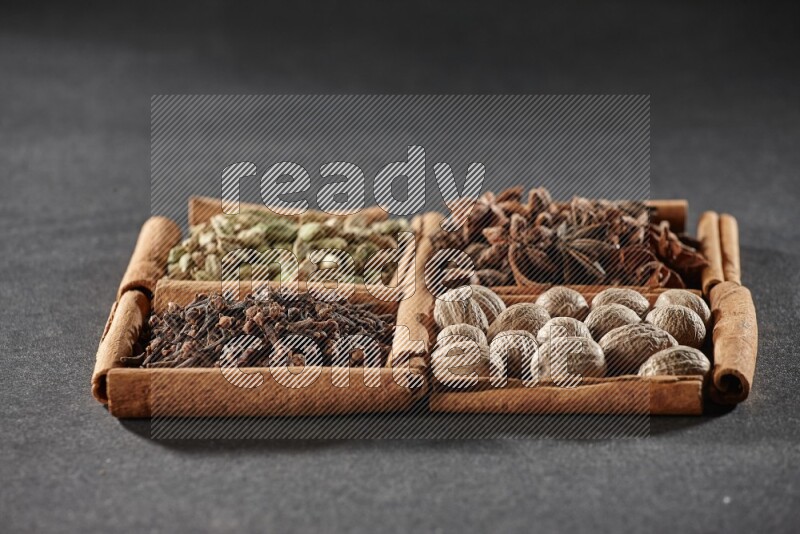 4 مربعات من عيدان القرفة ممتلئة بحب الهيل واليانسون النجمي وجوزة الطيب والقرنفل على أرضية سوداء