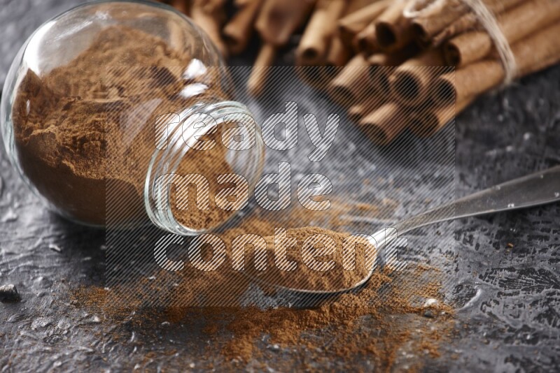 Herbal glass jar full cinnamon powder flipped and a metal spoon full of powder, cinnamon sticks stacked and bounded in the back on textured black background in different angles