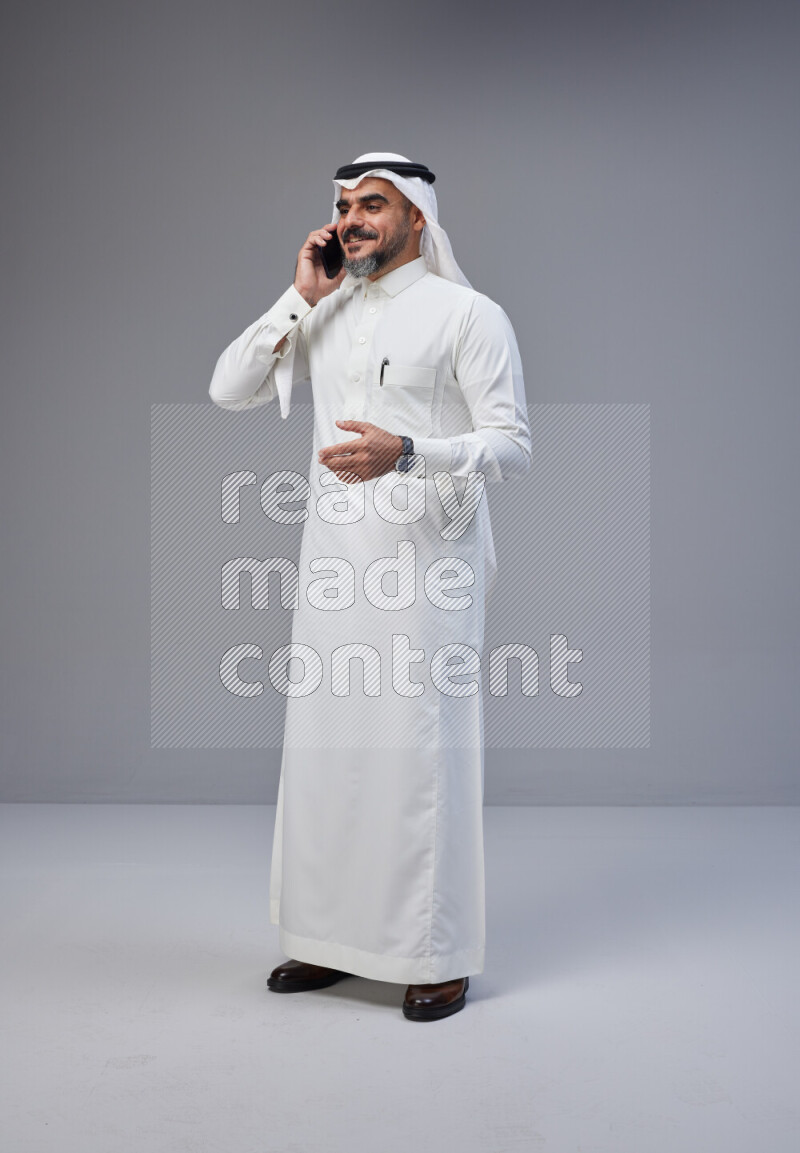 Saudi man Wearing Thob and white Shomag standing talking on phone on Gray background