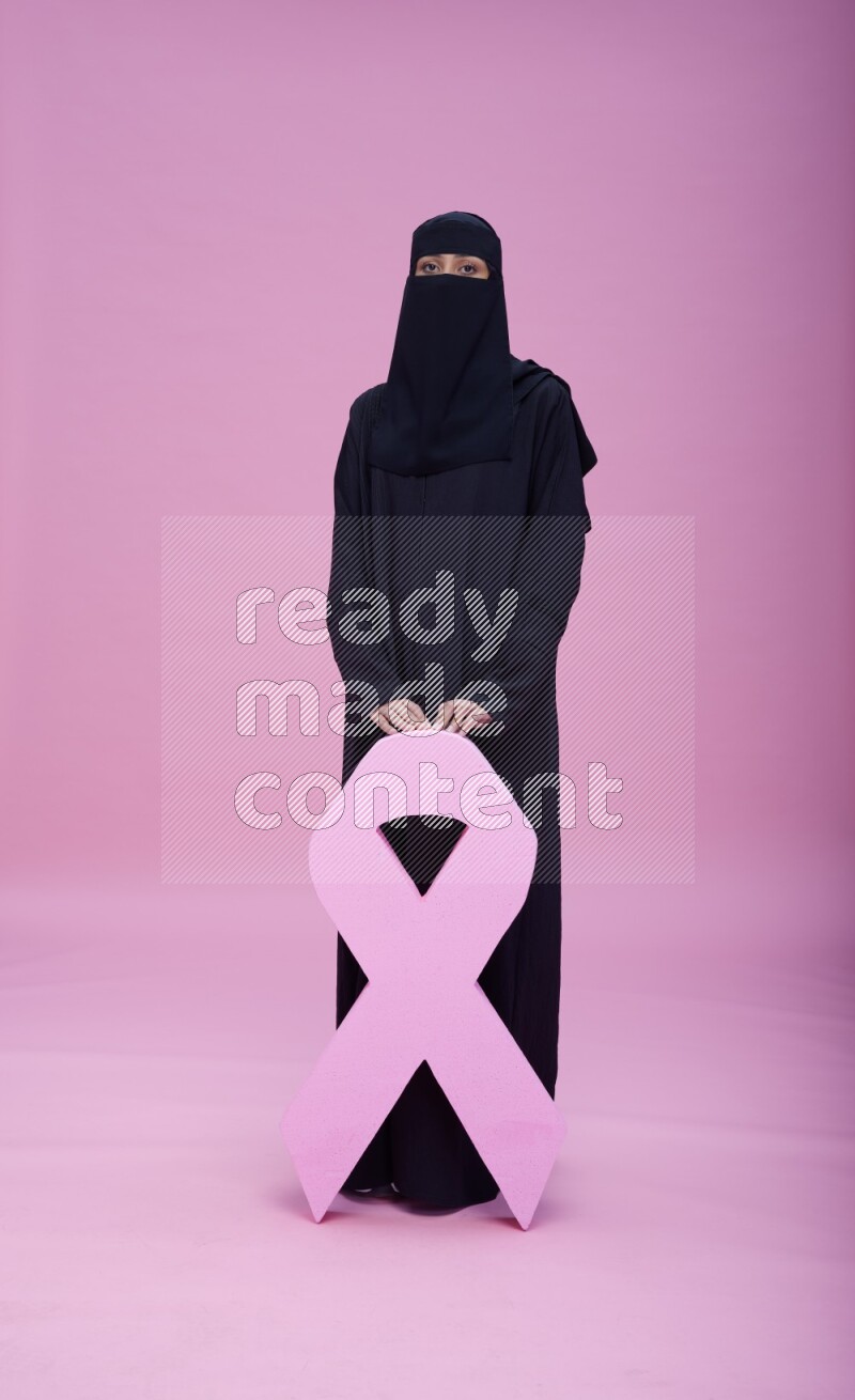 Saudi woman wearing abaya and niqab standing holding awareness ribbon on pink background