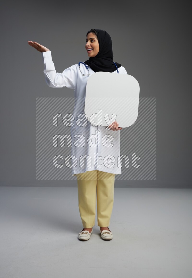 Saudi woman wearing lab coat with stethoscope standing holding social media sign on Gray background