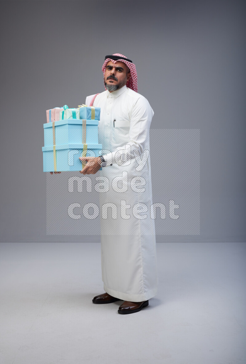 Saudi man Wearing Thob and red Shomag standing holding gift box on Gray background