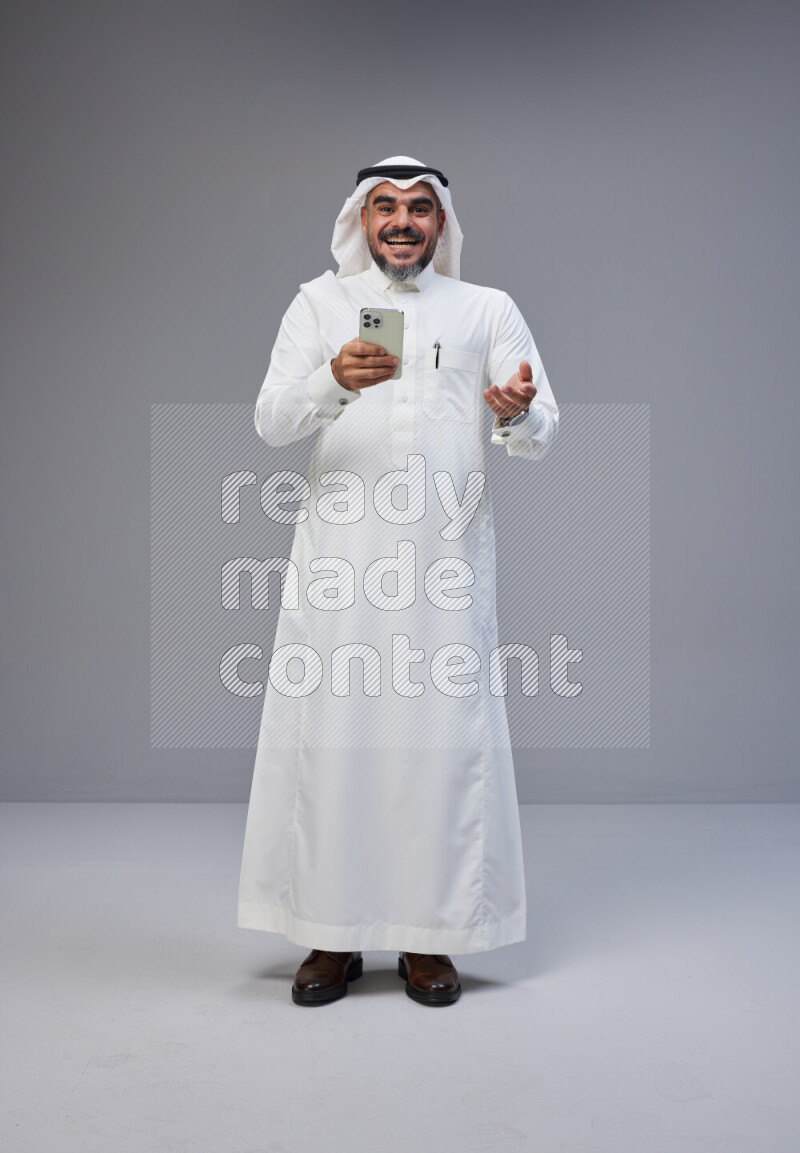 Saudi man Wearing Thob and white Shomag standing texting on phone on Gray background
