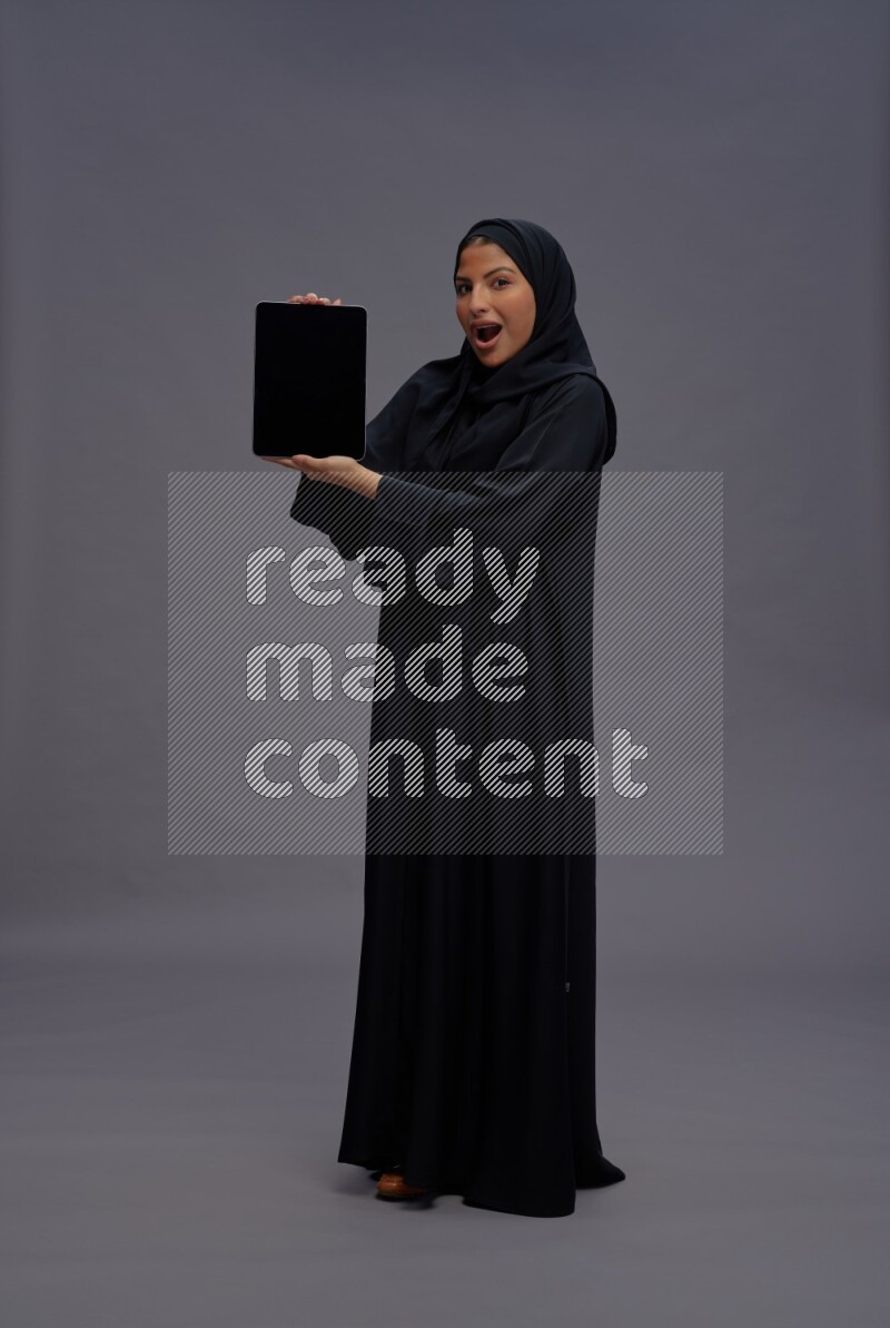 Saudi woman wearing Abaya standing showing tablet to camera on gray background
