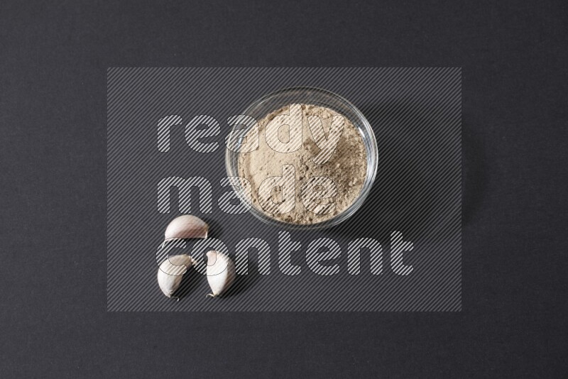 A glass bowl full of garlic powder with garlic bulb and some cloves beside it on a black flooring