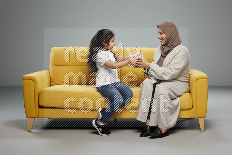 A girl sitting on a yellow sofa and giving a gift to her mother on gray background