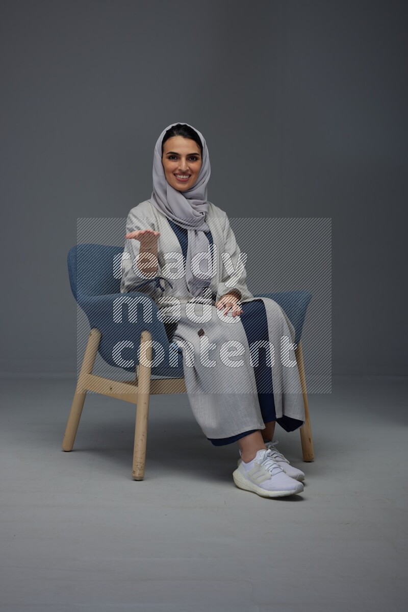 A Saudi woman wearing a light gray Abaya and head scarf sitting on a dark grey chair while making multi hand gestures eye level on a grey background