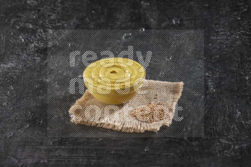 A glass bowl full of mustard paste set on a burlap piece with some mustard seeds on a textured black flooring