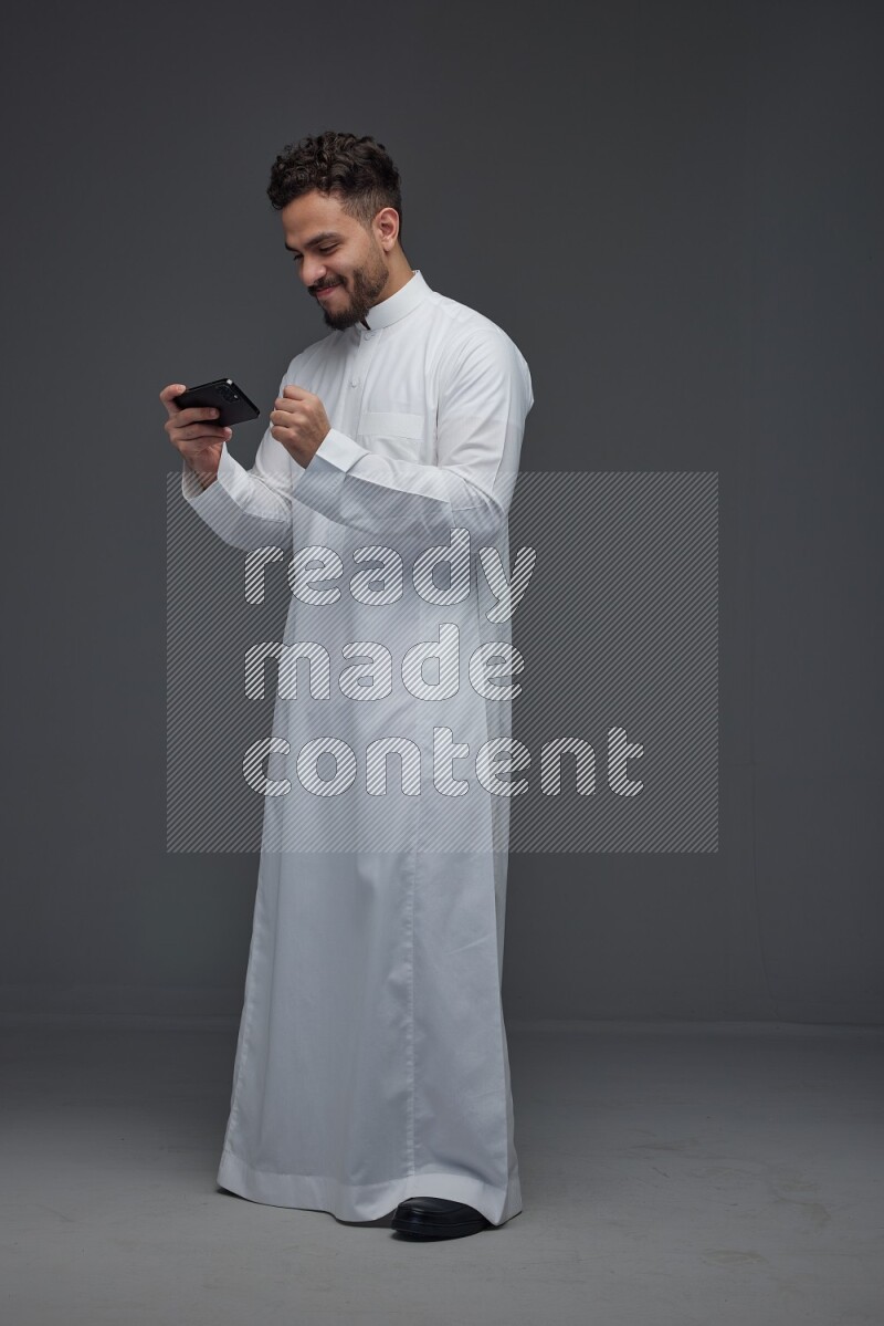 A Saudi man wearing Thobe and playing using his phone horizontally playing video games while standing and making different poses eye level on a gray background