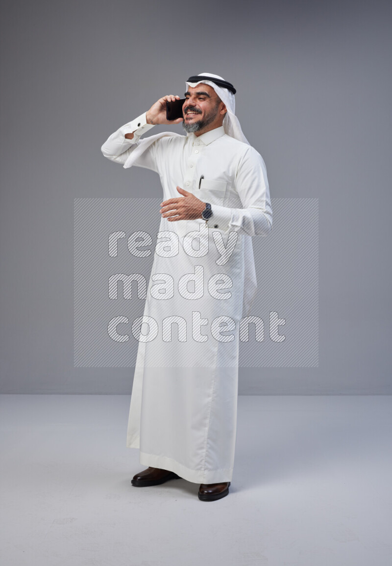 Saudi man Wearing Thob and white Shomag standing talking on phone on Gray background