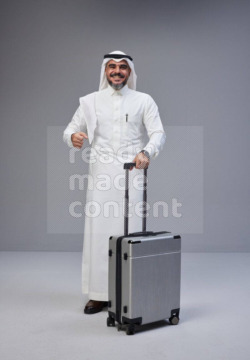 Saudi man wearing Thob and white Shomag standing holding Travel bag on Gray background