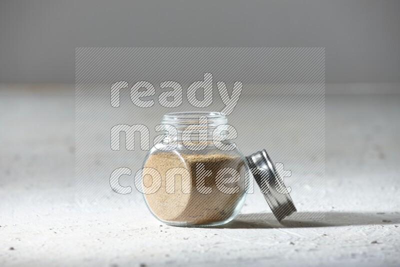 A glass spice jar full of cumin powder on textured white flooring