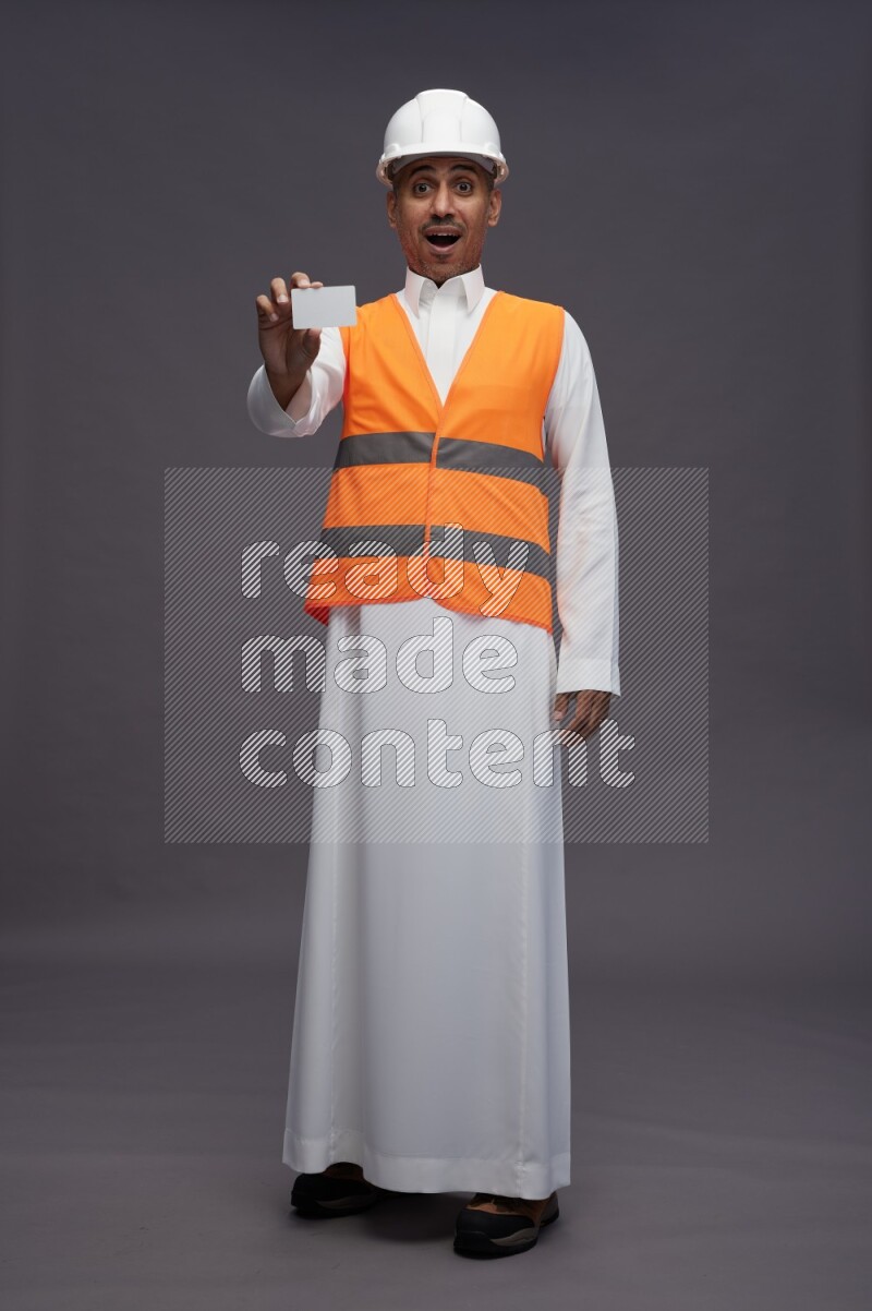 Saudi man wearing thob with engineer vest standing holding ATM card on gray background