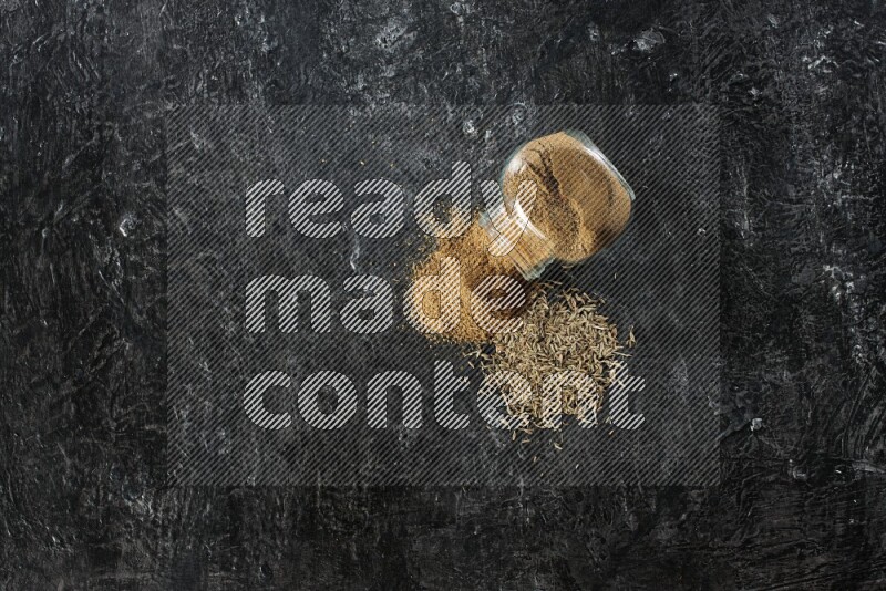 A flipped glass spice jar full of cumin powder with spilled powder and cumin seeds on a textured black flooring