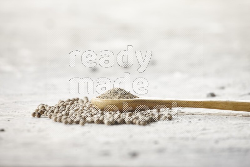 A wooden spoon full of white pepper powder and white pepper beads on textured white flooring