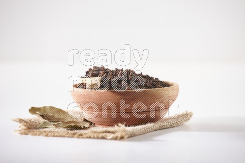 A wooden bowl and a wooden spoon full of cloves on a piece of burlap on a white flooring