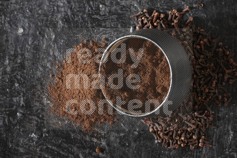 A black pottery bowl full of cloves powder spilled out of it with whole grains on a textured black flooring