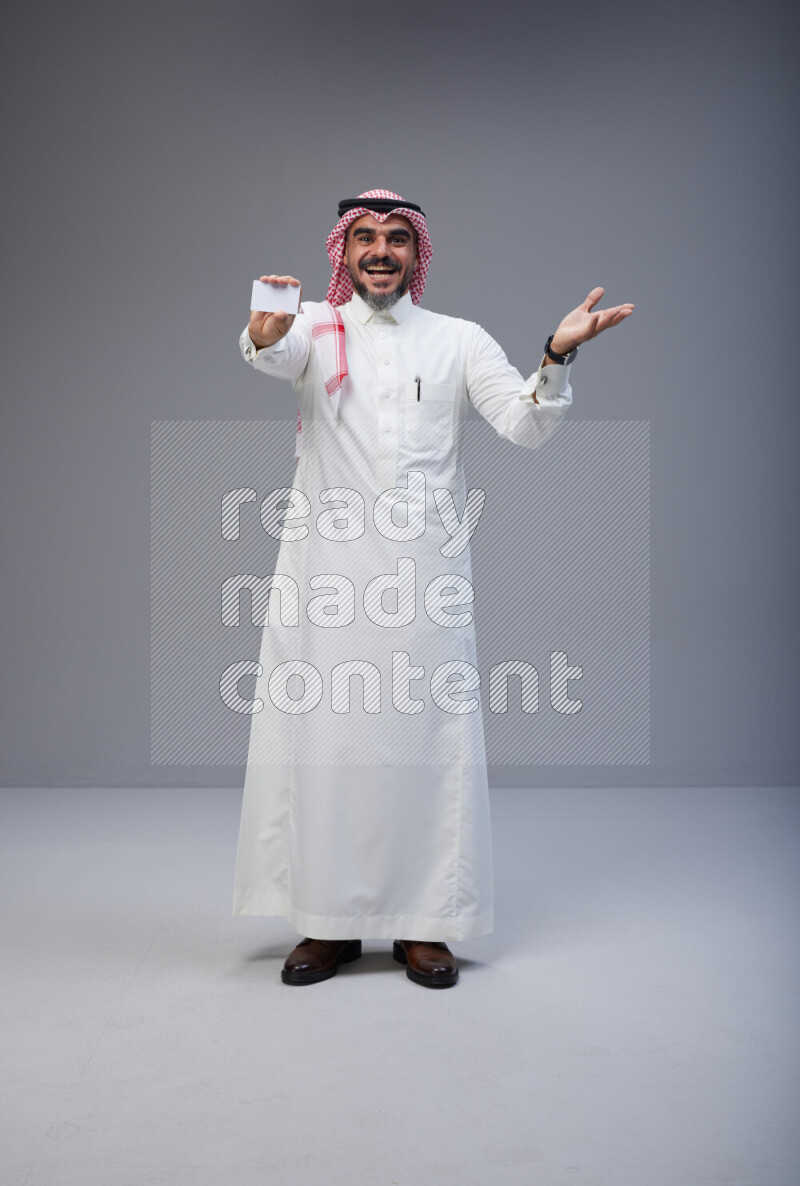 Saudi man Wearing Thob and red Shomag standing holding ATM card on Gray background