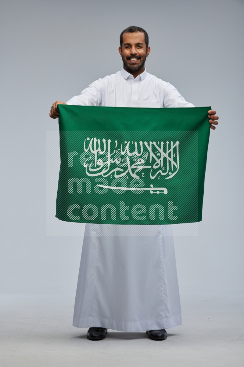 Saudi man wearing Thob standing holding Saudi flag on Gray background