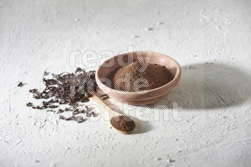 A wooden bowl and wooden spoon full of cloves powder with cloves spread on textured white flooring