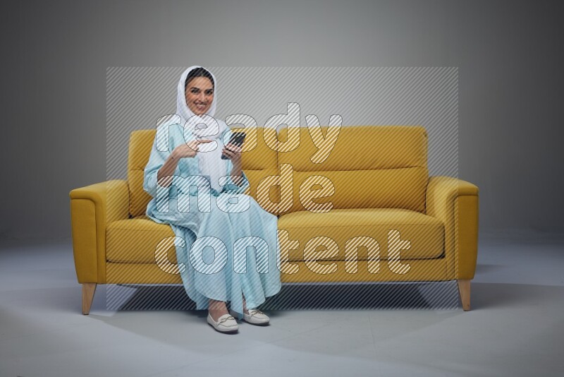 A Saudi woman wearing a light blue Abaya and a white head scarf sitting on a yellow sofa and holding her phone while pointing to it eye level on a grey background