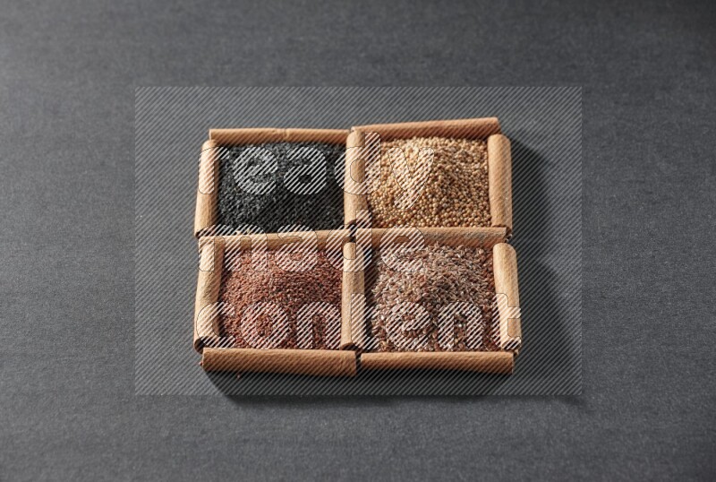 4 مربعات من عيدان القرفة ممتلئة بذور الكتان وحب الرشاد وحبة البركة السوداء وحبوب الخردل على أرضية سوداء