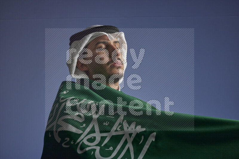 A close-up shot of Saudi man wearing thob and white shomag wrapping big Saudi flag on gray background