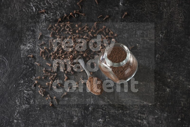 A glass spice jar and a metal spoon full of cloves powder with cloves spread on textured black flooring