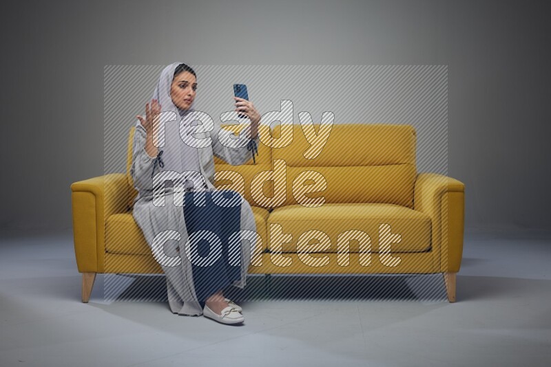 A Saudi woman wearing a light gray Abaya and head scarf sitting on a yellow sofa and making a video call eye level on a grey background