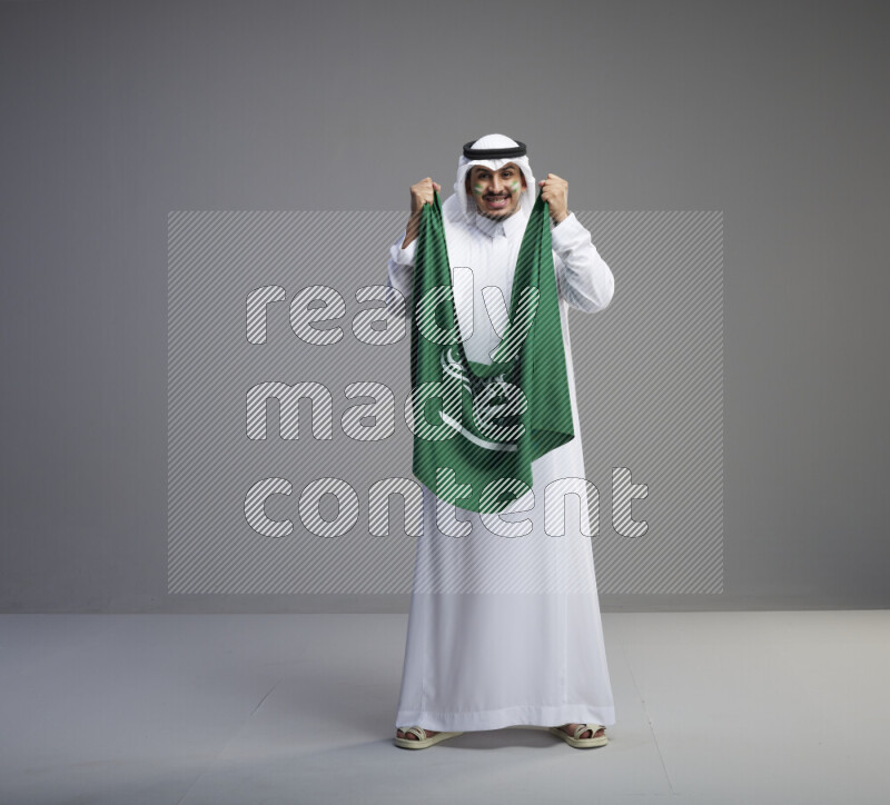 A Saudi man standing wearing thob and white shomag with face painting holding big Saudi flag on gray background