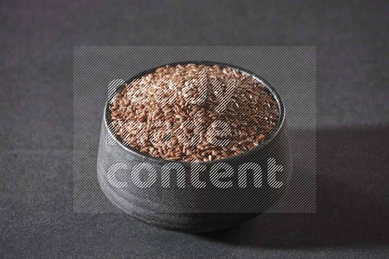 A black pottery bowl full of flaxseeds on a black flooring