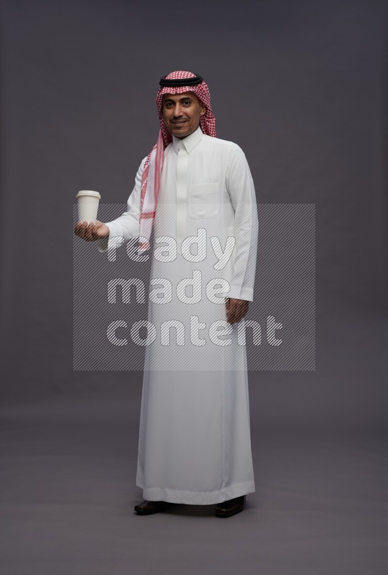 Saudi man wearing thob and shomag standing holding paper cup on gray background
