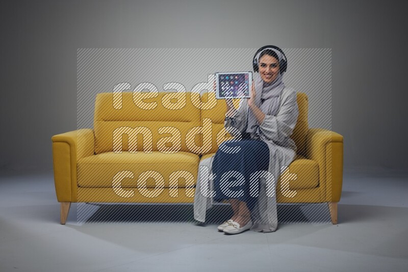 A Saudi woman wearing a light gray Abaya and head scarf sitting on a yellow sofa and using her phone while wearing headphones eye level on a grey background
