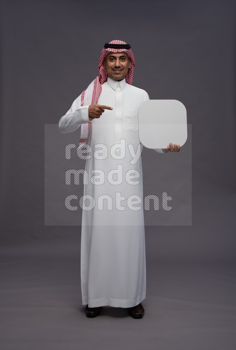 Saudi man wearing thob and shomag standing holding social media sign on gray background