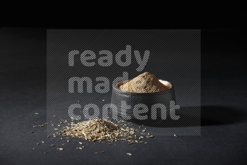 A black pottery bowl full of cumin powder and spreaded cumin seeds on a black flooring