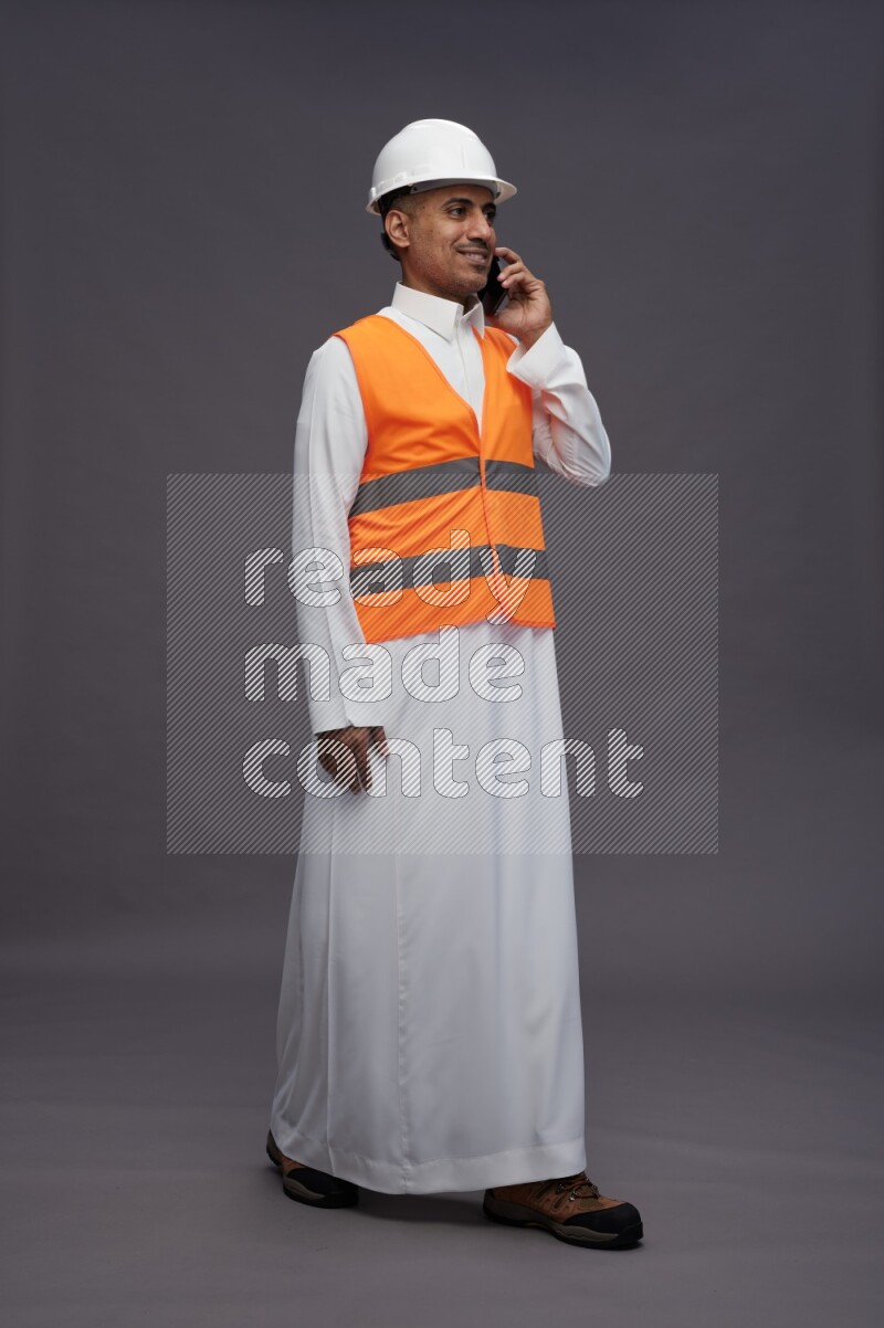 Saudi man wearing thob with engineer vest standing talking on phone on gray background