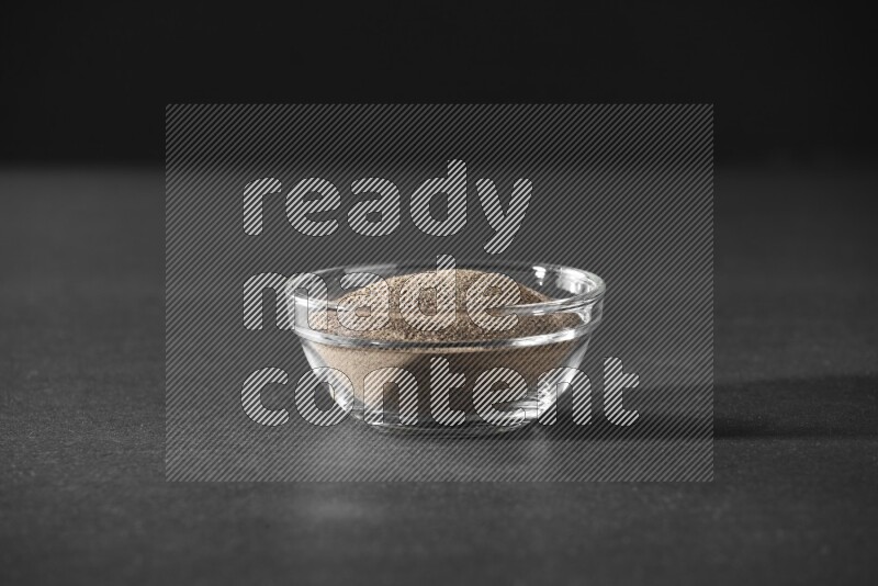 A glass bowl full of black pepper powder on black flooring