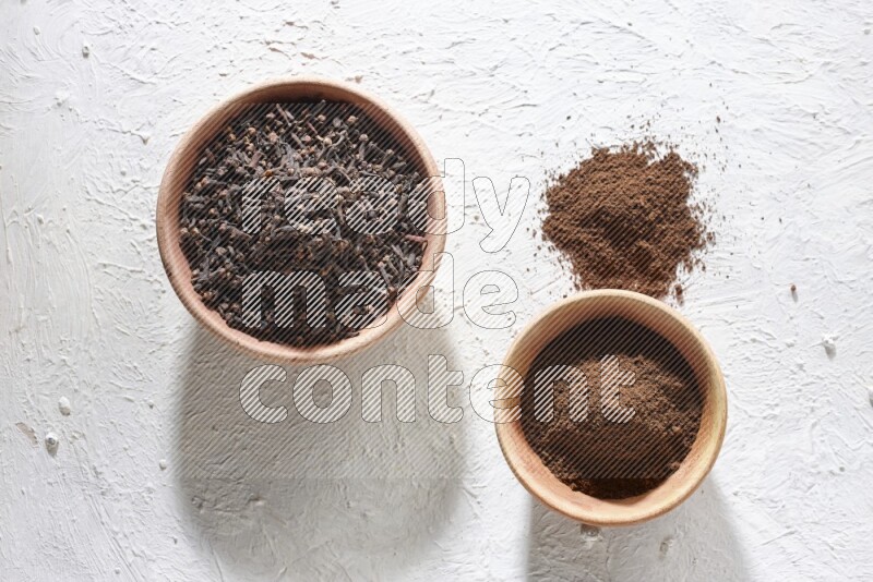 2 wooden bowls full of cloves powder and whole cloves on a textured white flooring