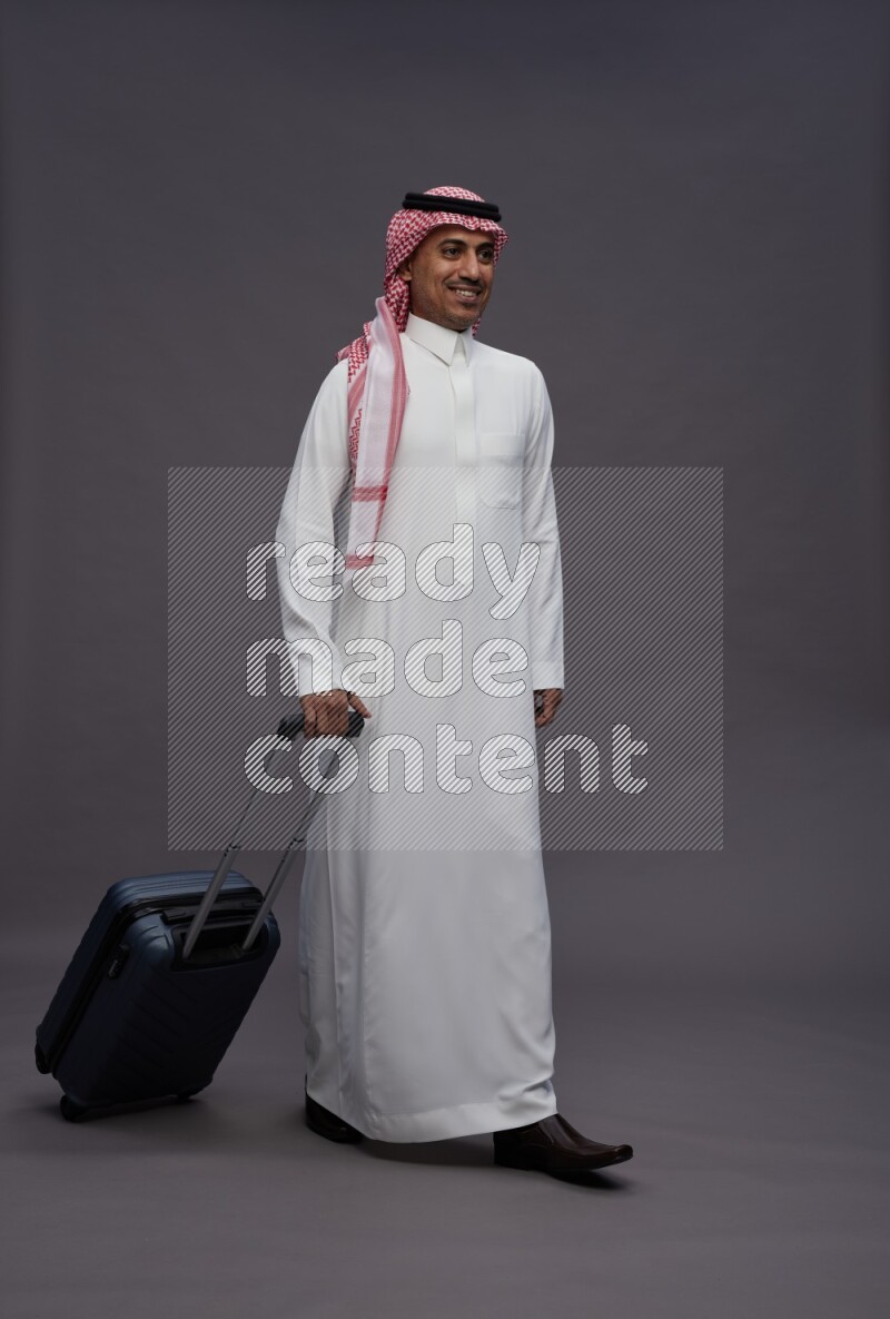 Saudi man wearing thob and shomag standing holding bag on gray background