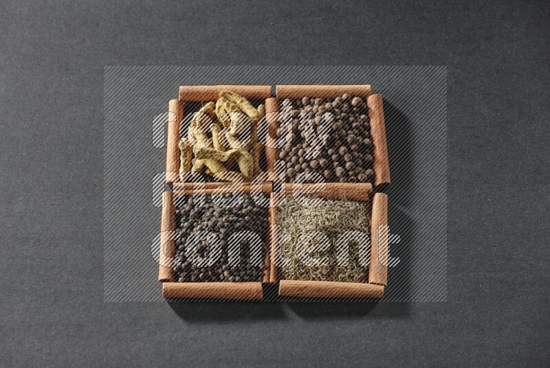 4 مربعات من عيدان القرفة ممتلئة بحبوب الفلفل الأسود والكمون وبهار الفلفل الحلو وأصابع الكركم على أرضية سوداء